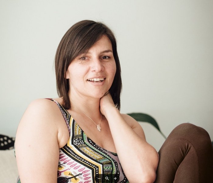 Manuela die auf einem Stuhl sitzt, eine Hand hält sie am Hals. Sie lächelt verschmitzt in die Kamera