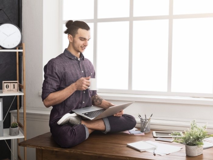 Mann sitzt im Yoga-Schneidersitz mit Laptop und einer Tasse auf seinem Schreibtisch.